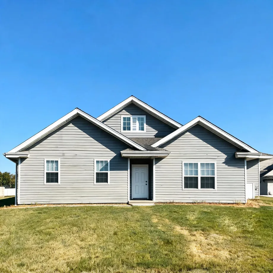 Single-story gray horizontal-siding house with multiple front gables, a white front door, white-trimmed windows, and a mown lawn under a clear blue sky.