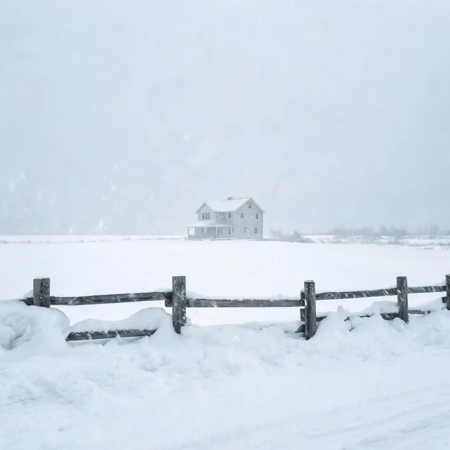 A two-story house with a porch stands in a snow-covered field under falling snow, with a weathered wooden rail fence partly buried in drifts in the foreground.