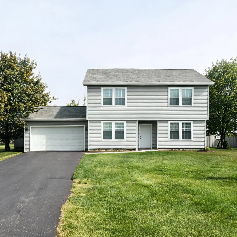 A two-story light gray vinyl-sided house with an attached two-car garage, asphalt driveway, mown lawn, and deciduous trees on either side under a pale sky.