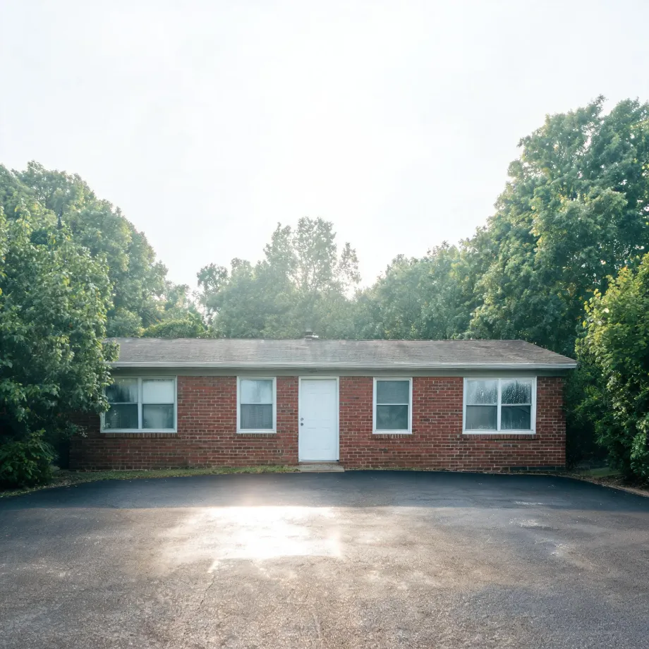 A single-story red brick house with a white door and white-framed windows, backed by dense green trees and fronted by a large asphalt parking area under hazy bright sky.