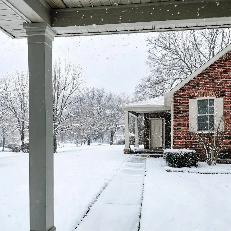 View from under a covered porch with a white column toward a brick house with a white front door and shuttered window, with snow falling and covering the lawn, walkway, shrubs, and bare trees.