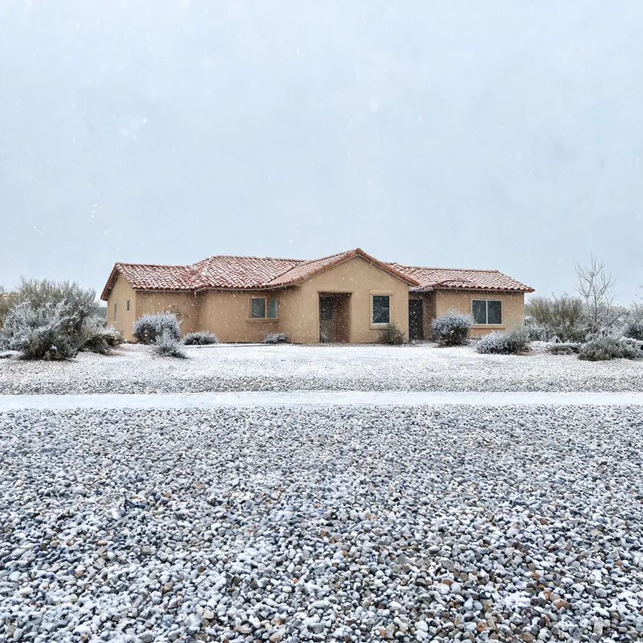 A single-story tan stucco house with a red tile roof behind a gravel yard and low desert shrubs, all lightly covered in snow under an overcast sky.