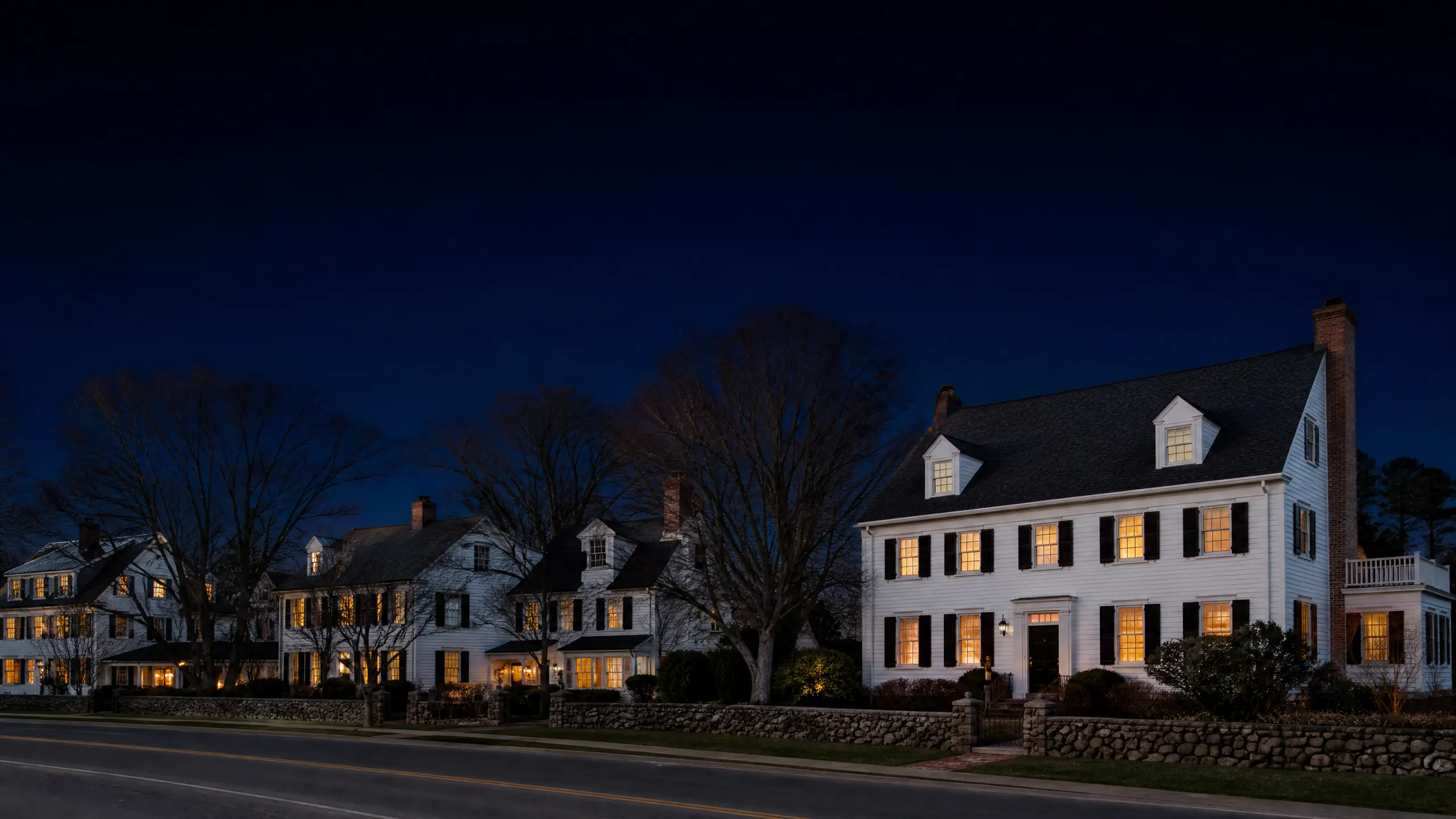 A row of illuminated white colonial houses along a road photographed at dusk.