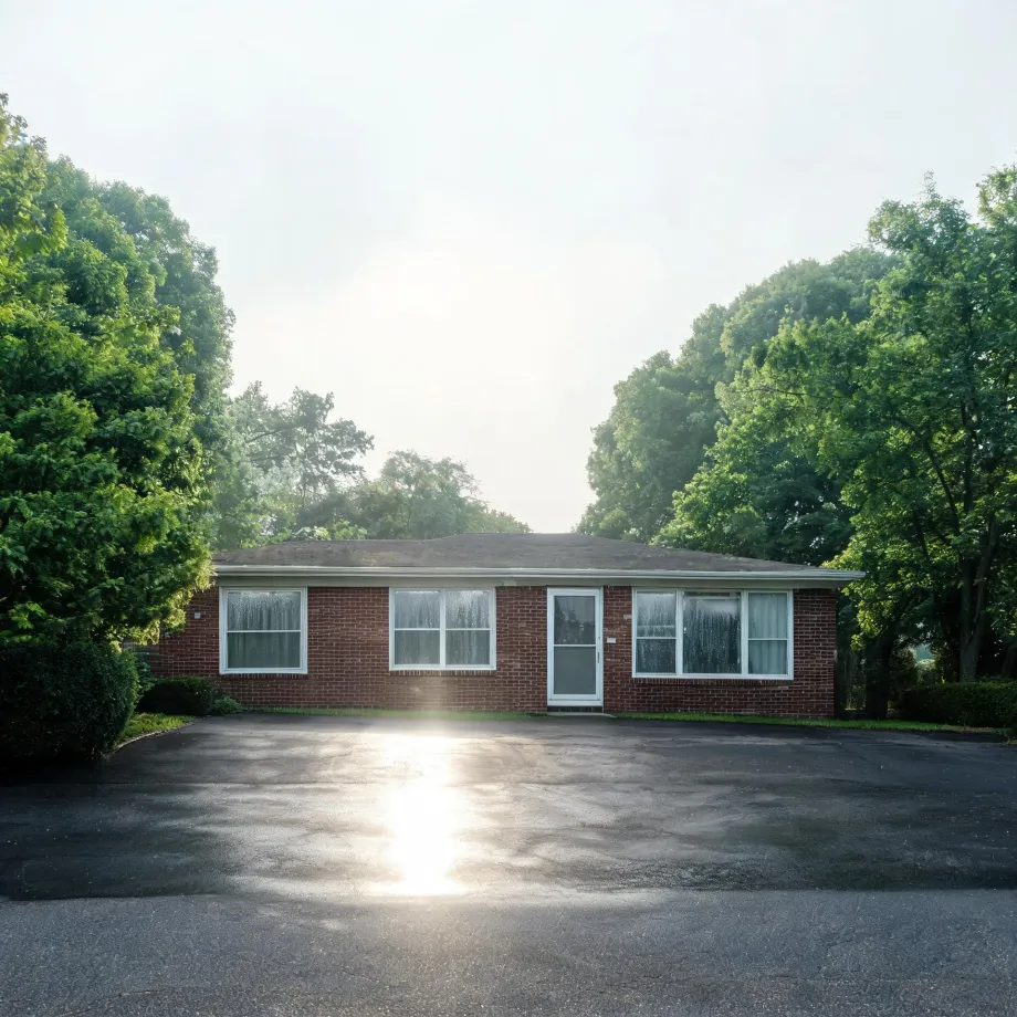 A single-story red brick house with white-framed windows and a white storm door, fronted by a large asphalt parking area with sun glare, surrounded by green deciduous trees and shrubs under a bright hazy sky.