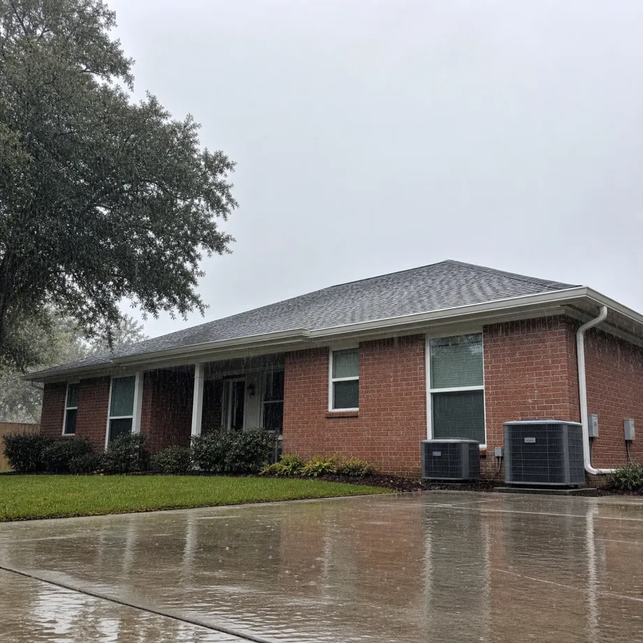 Single-story red brick house with a gray shingled hip roof, two outdoor air conditioning condenser units along the side, shrubs and lawn in front, and a wet concrete driveway during rain under an overcast sky.