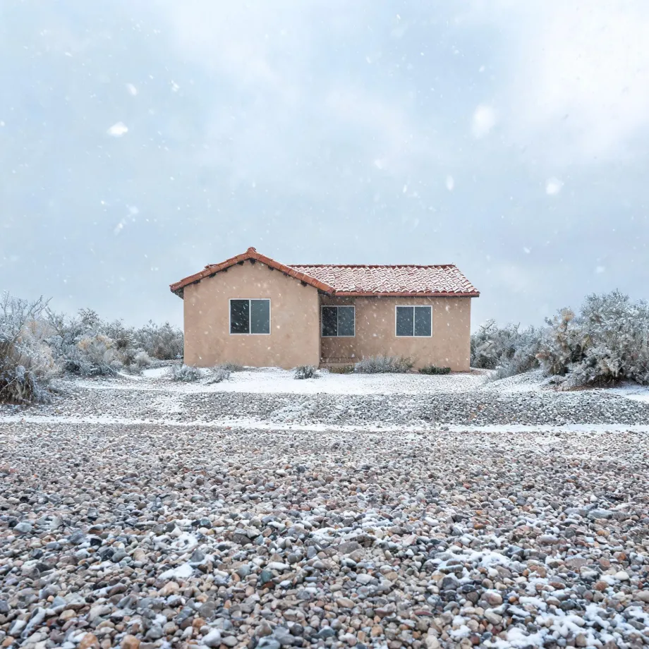 A single-story tan stucco house with a red tile roof stands behind a broad gravel lot dusted with snow, flanked by frost-covered desert shrubs under a pale sky with falling snowflakes.