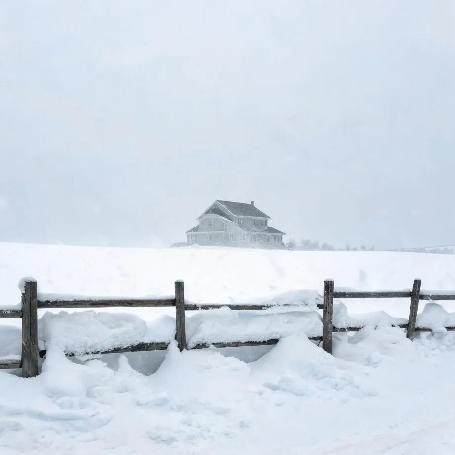 A two-story gabled house stands on a snow-covered rise under a pale overcast sky, with a snow-laden wooden rail fence crossing the foreground.