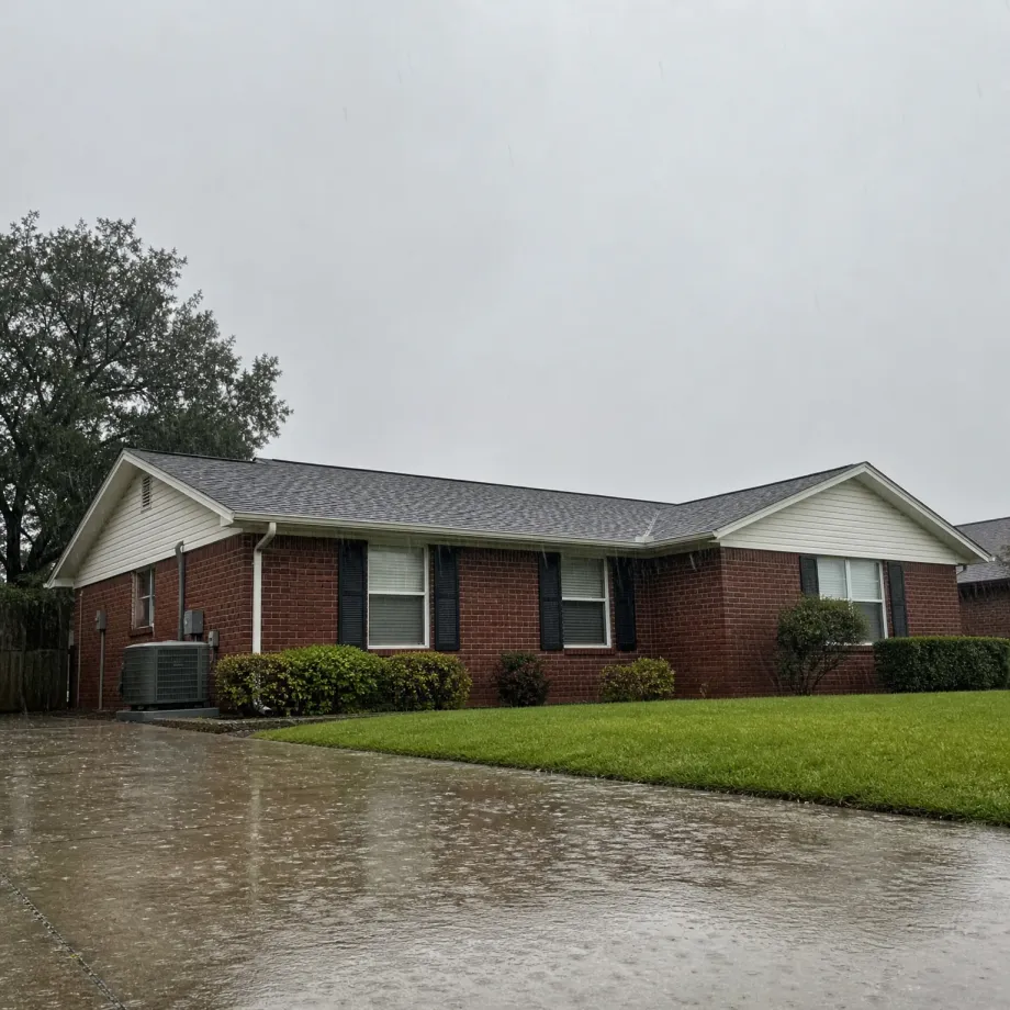 Single-story red brick house with gray shingled roof, white gable siding, black shutters and trimmed hedges, seen during rainfall with a wet driveway in the foreground and green lawn to the right.
