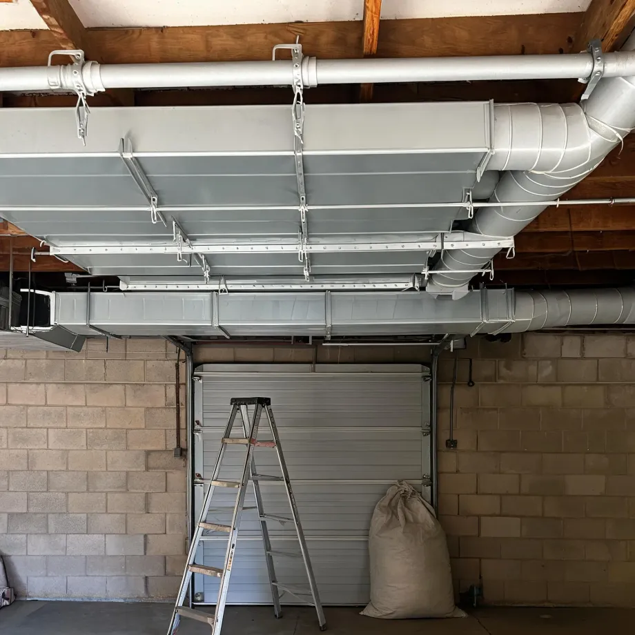 Interior of a garage with large rectangular and round sheet-metal ducts suspended from wooden ceiling joists, a step ladder and a filled sack in front of a sectional garage door in a concrete block wall.