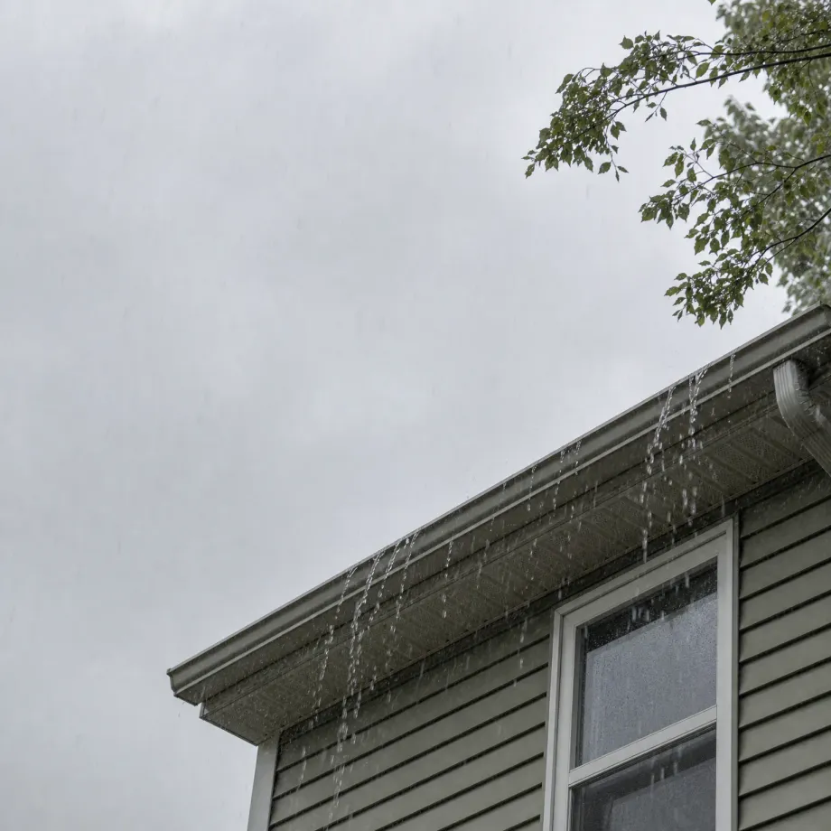 Water overflows from a gutter along the roof edge of a house with light horizontal siding and a white-framed window, seen from below against an overcast sky with tree branches at upper right.