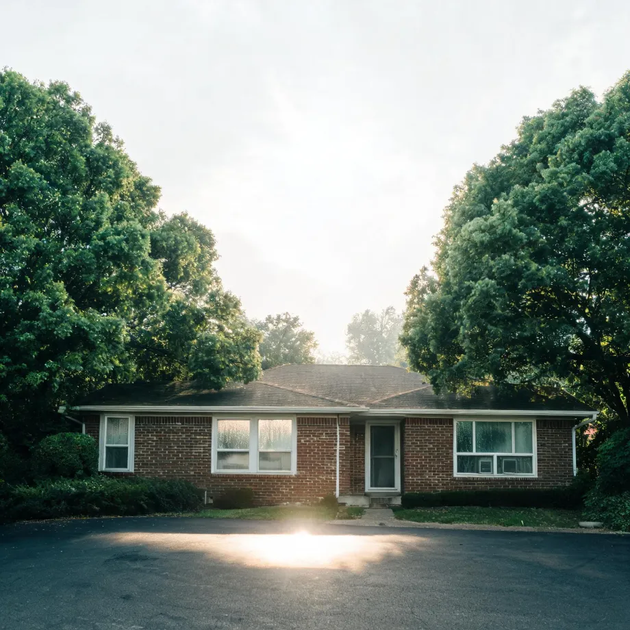 A single-story red brick ranch house with a low hipped roof, large picture windows and a central door, flanked by large leafy trees, with an empty asphalt lot in the foreground lit by low sun flare.