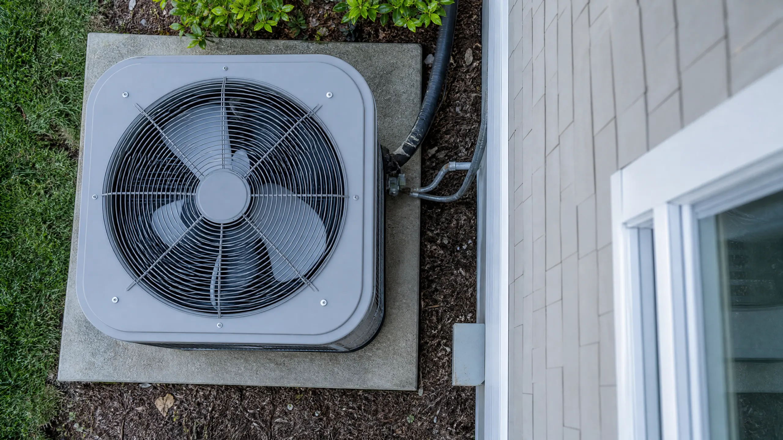 Aerial view of a residential air conditioning condenser unit mounted on a concrete pad next to a house with vinyl siding and a sliding glass door.
