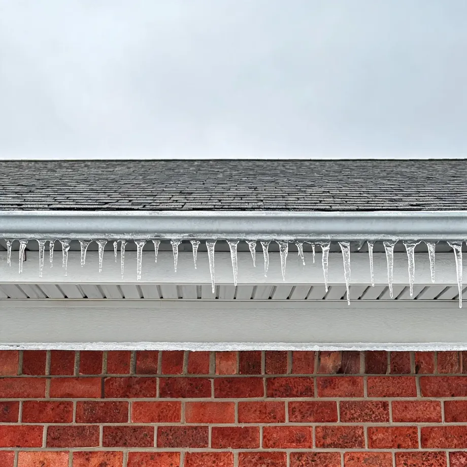 Row of small icicles hanging from a white gutter along a shingled roof edge, above a vented soffit and red brick wall, under an overcast sky.