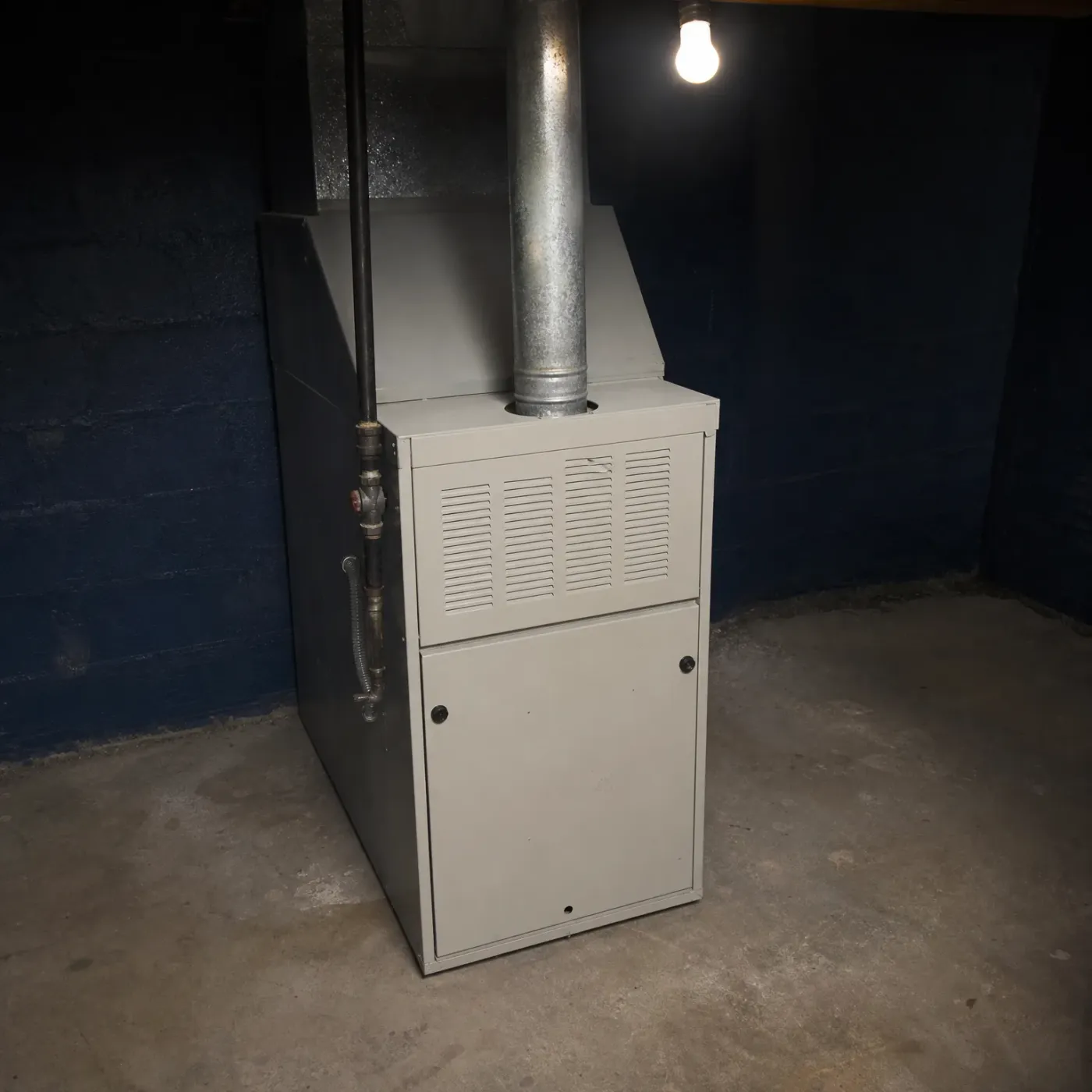 A beige residential furnace with a louvered upper panel stands on a concrete basement floor beside a dark blue painted block wall, with a galvanized metal flue pipe rising from its top and a bare lit bulb overhead.