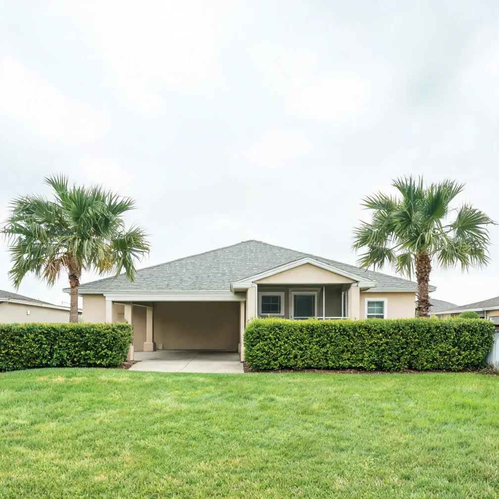 A single-story beige stucco house with a gray shingled hip roof and an open carport, flanked by two palm trees and a trimmed hedge, with a mown lawn in the foreground under an overcast sky.