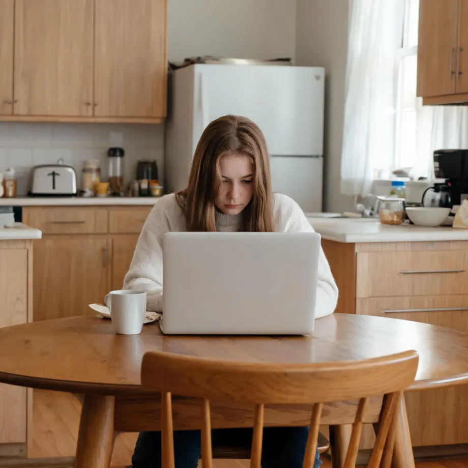 A young woman in a cream sweater sits at a round wooden table using a white laptop, with a white mug beside her in a kitchen with wood cabinets and a white refrigerator.