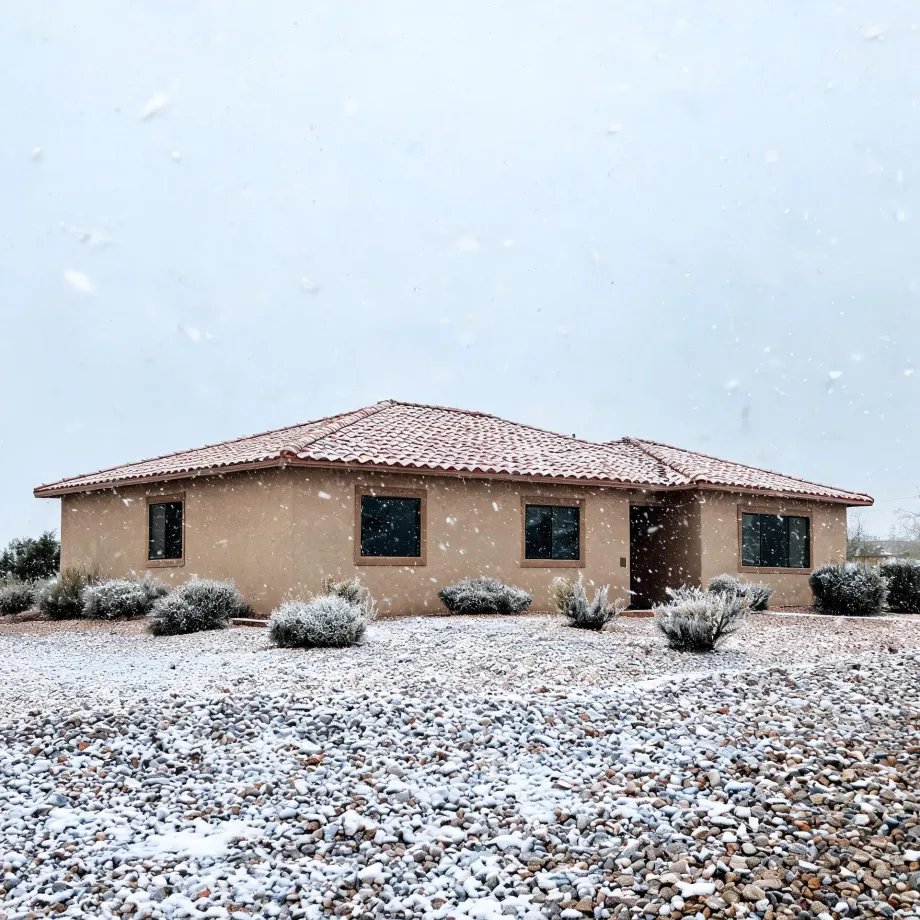 A single-story tan stucco house with a red tile roof during falling snow, fronted by a gravel yard and low shrubs dusted with snow under an overcast sky.