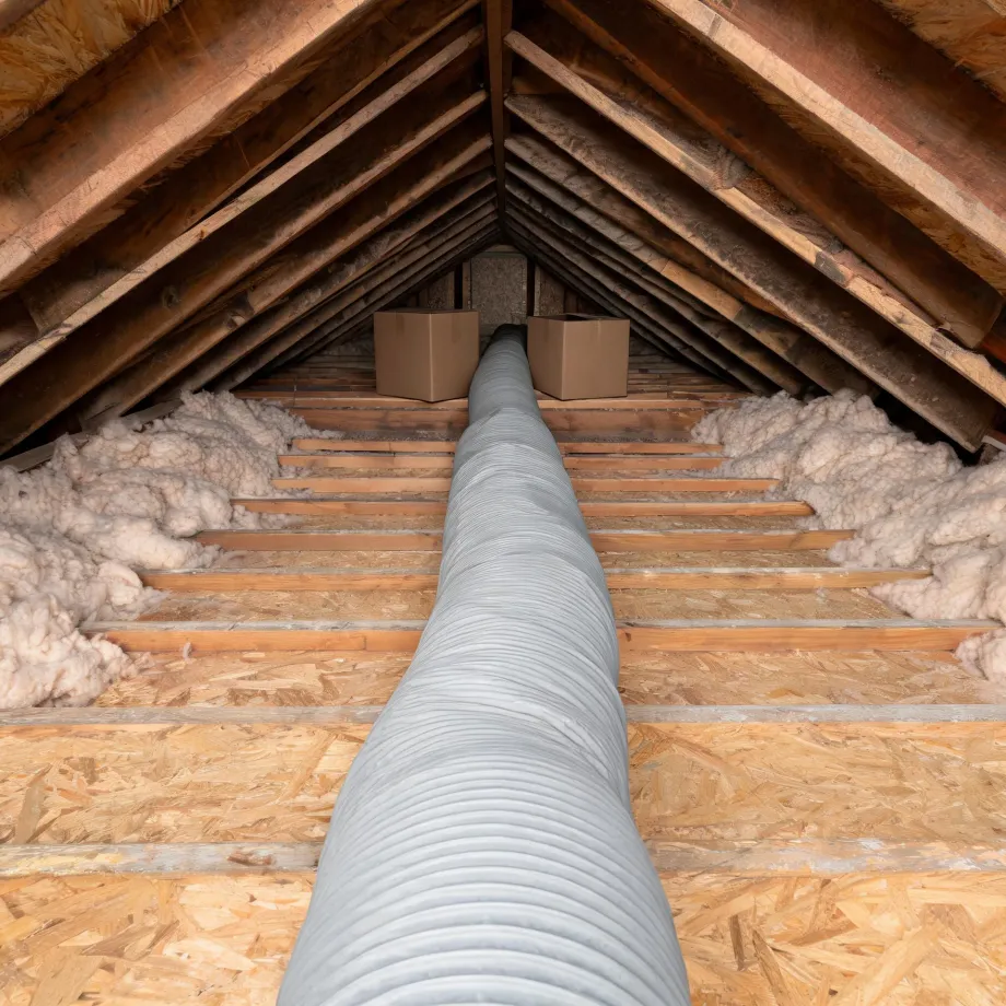 Attic interior with exposed wooden rafters, an OSB floor, batts of insulation along the eaves, two cardboard boxes at the far end, and a flexible gray duct running down the center.
