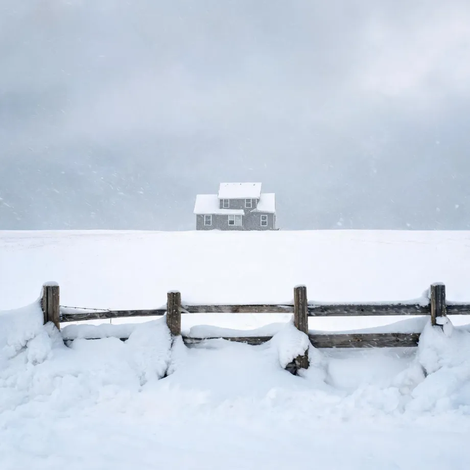 A gray two-story house with snow-covered roofs stands on a snow-covered rise under an overcast sky, with a snow-laden wooden post-and-rail fence in the foreground and falling snow.
