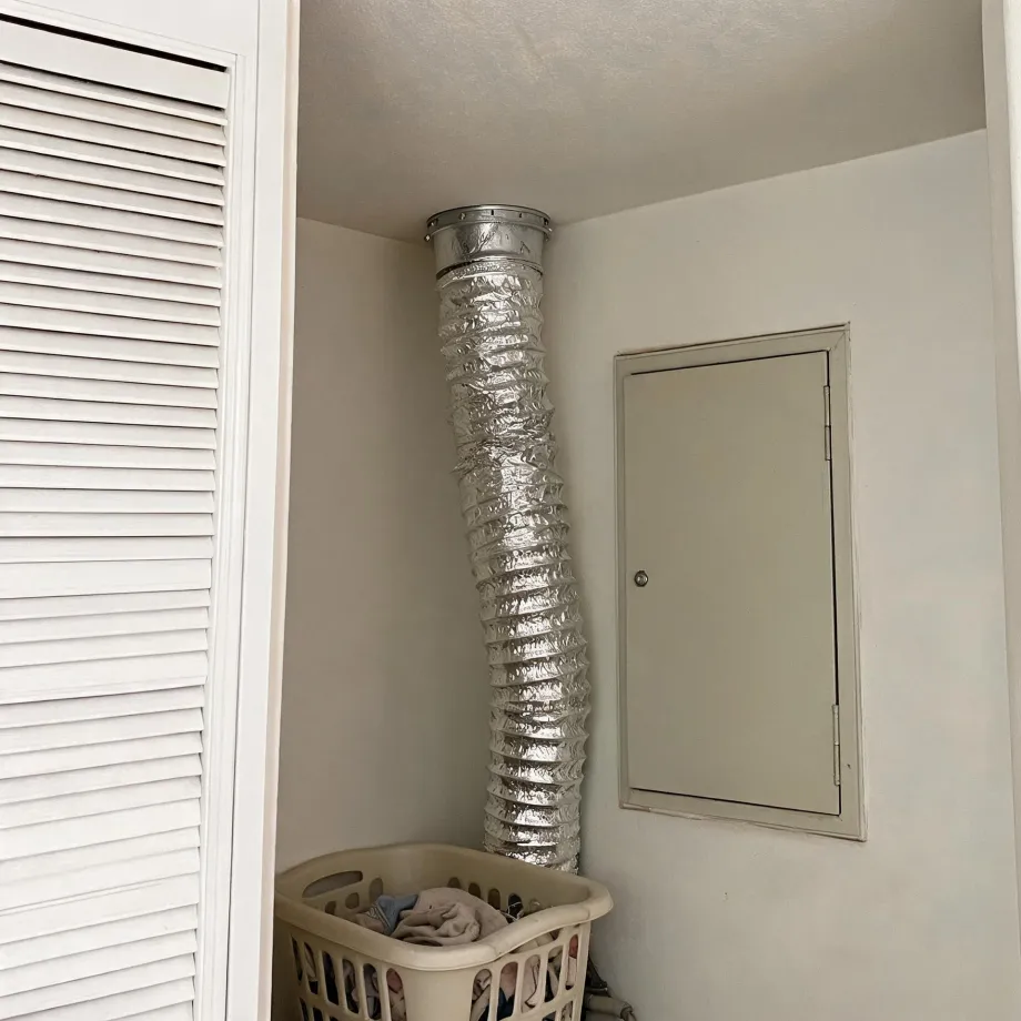 Interior corner of a laundry closet where a flexible foil dryer duct runs from the floor up to a metal collar in the ceiling, next to a louvered white door and a square wall access panel, with a plastic laundry basket of clothes on the floor.