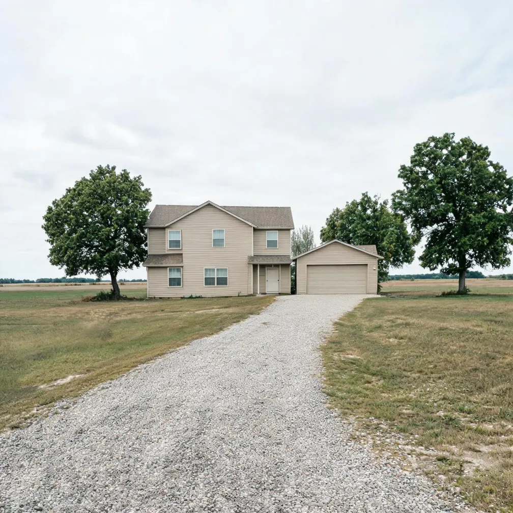 A two-story beige vinyl-sided house with an attached two-car garage sits in flat open grassland with two large deciduous trees, approached by a wide gravel driveway under an overcast sky.