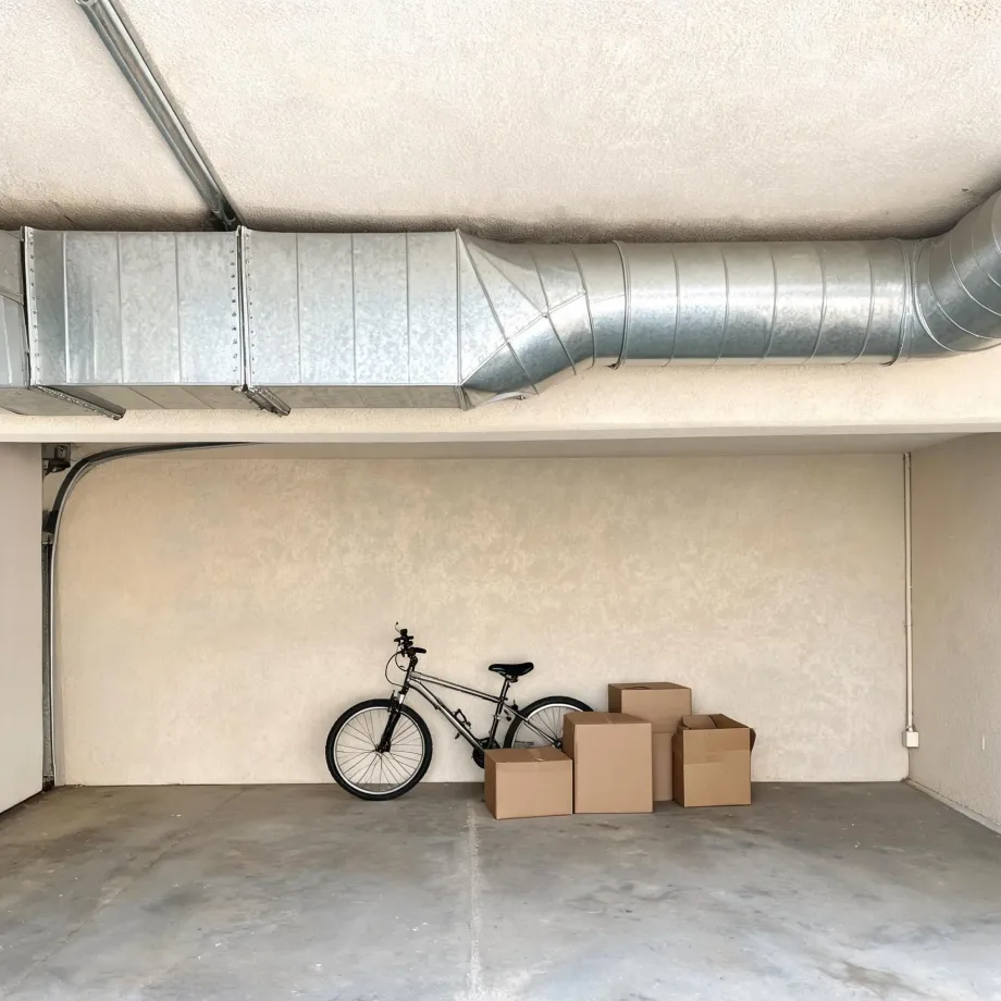 Interior of an empty garage with a concrete floor, a bicycle leaning against a textured beige wall beside three cardboard boxes, and exposed galvanized metal ductwork running along the ceiling.