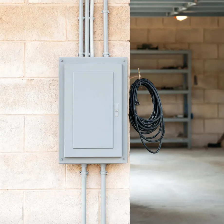 A gray electrical panel box mounted on a concrete block wall with conduit pipes running above and below it, and a coiled black extension cord hanging on the wall beside an open garage area with metal shelving.