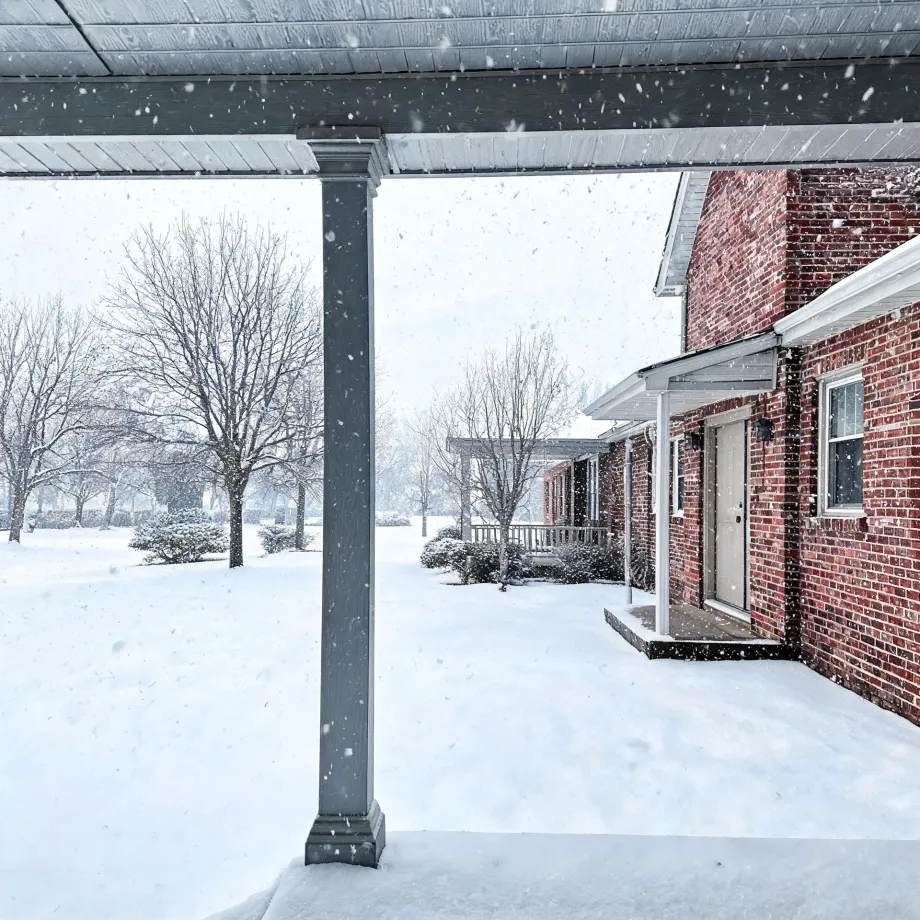 View from under a covered porch with a square gray column toward a red brick house with a white door and small roof overhang, with falling snow, snow-covered ground, and bare trees in the background.