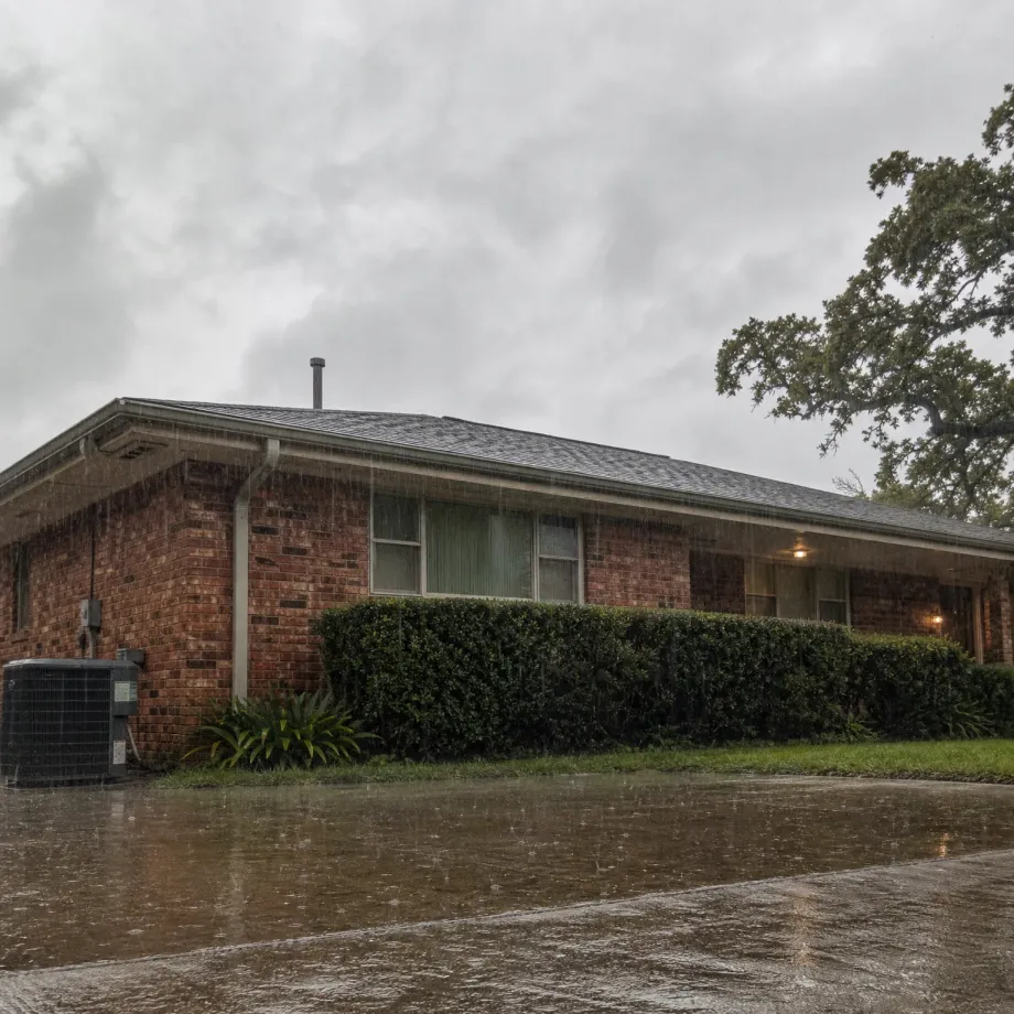 A single-story brick house with a low-pitched shingled roof, trimmed hedges and an outdoor air conditioning unit, seen during rainfall with a wet driveway in the foreground and an overcast sky.