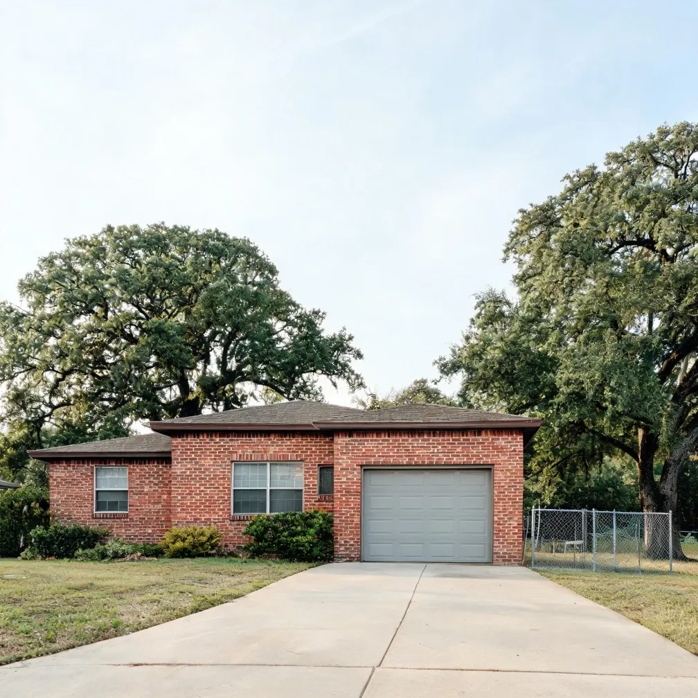Single-story red brick house with a white single-car garage door, a concrete driveway leading to the viewer, shrubs along the front wall, a chain-link fence at right, and large leafy trees behind.