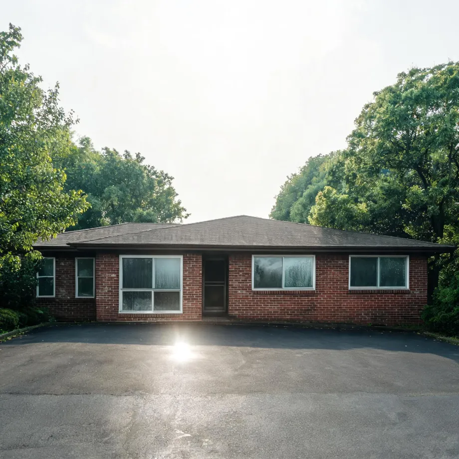 A single-story red brick building with a shingled hip roof, large windows and a recessed central entry, fronted by a wide empty asphalt parking area with sun glare, surrounded by green deciduous trees.