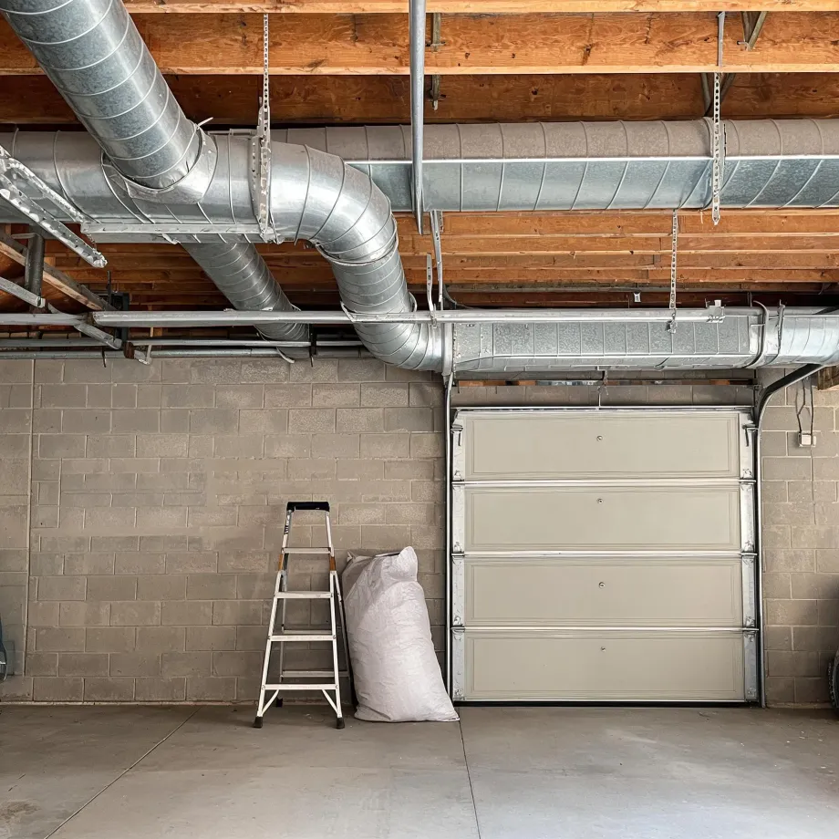 Garage interior with exposed galvanized round and rectangular ductwork under wood joists, concrete block walls, a closed paneled overhead door, a folding step ladder, and a white sack on the concrete floor.