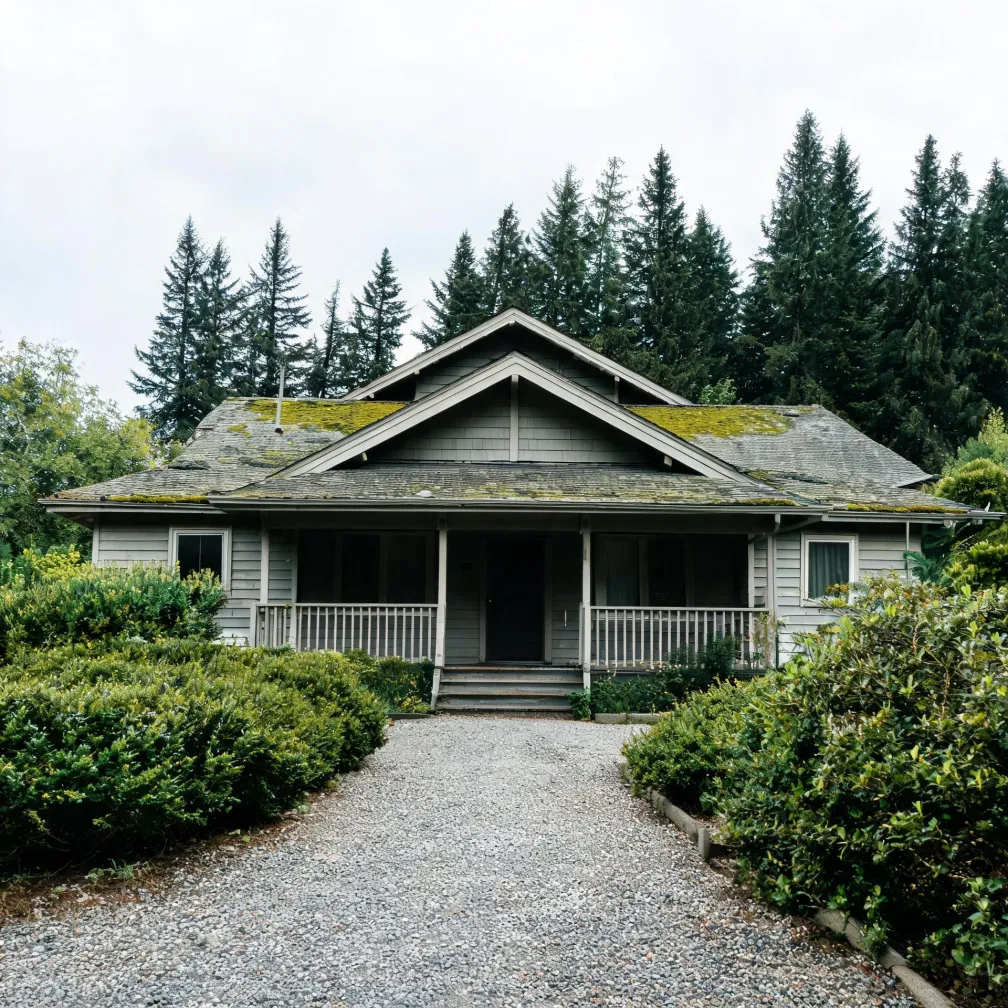 A single-story gray shingled house with a covered porch and moss-covered roof, fronted by a gravel driveway and trimmed hedges, with tall evergreen trees behind it under an overcast sky.