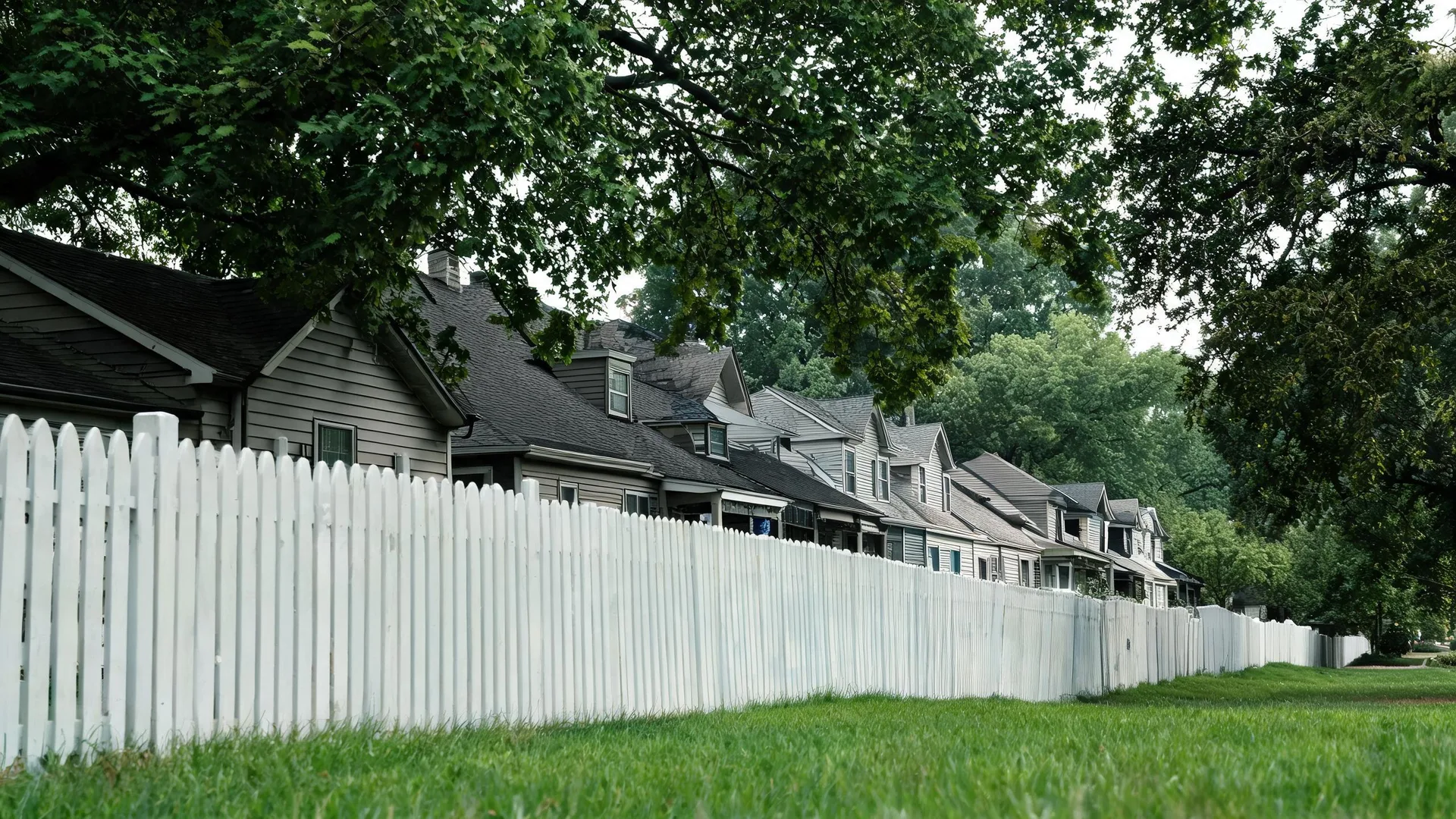 A long white picket fence runs beside a mown grass strip in front of a row of gray gabled houses, with large leafy tree branches overhead.