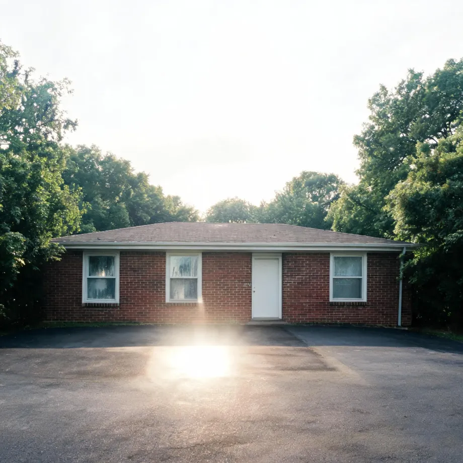 A single-story red brick house with a white door and three white-framed windows, fronted by an asphalt parking area with sun flare, surrounded by green deciduous trees under a bright hazy sky.