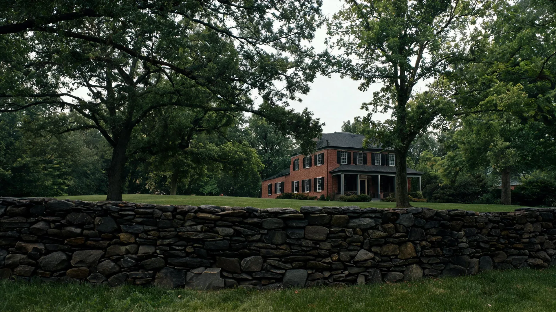 A two-story brick house with black shutters and a covered porch stands on a mown lawn behind large deciduous trees, with a dry-stacked stone wall running across the foreground.