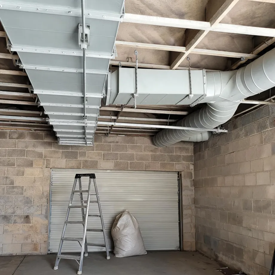 Interior of a concrete block garage with rectangular and round sheet-metal ductwork suspended from the timber ceiling joists, a step ladder and a filled sack standing in front of a closed roller shutter door.