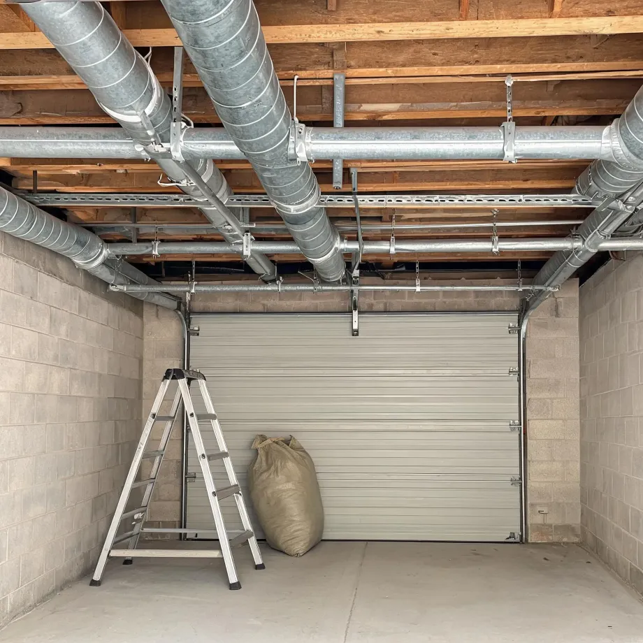 Interior of a concrete block garage with a closed white roll-up sectional door, an aluminum step ladder and a burlap sack on the floor, and exposed wooden joists with galvanized ducts and conduit overhead.