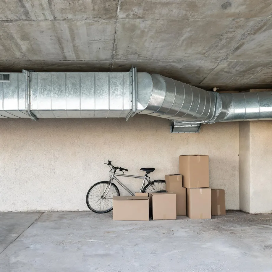 A bicycle leaning against a beige stucco wall beside several cardboard boxes in a concrete-ceilinged garage, with large galvanized metal ductwork running overhead.
