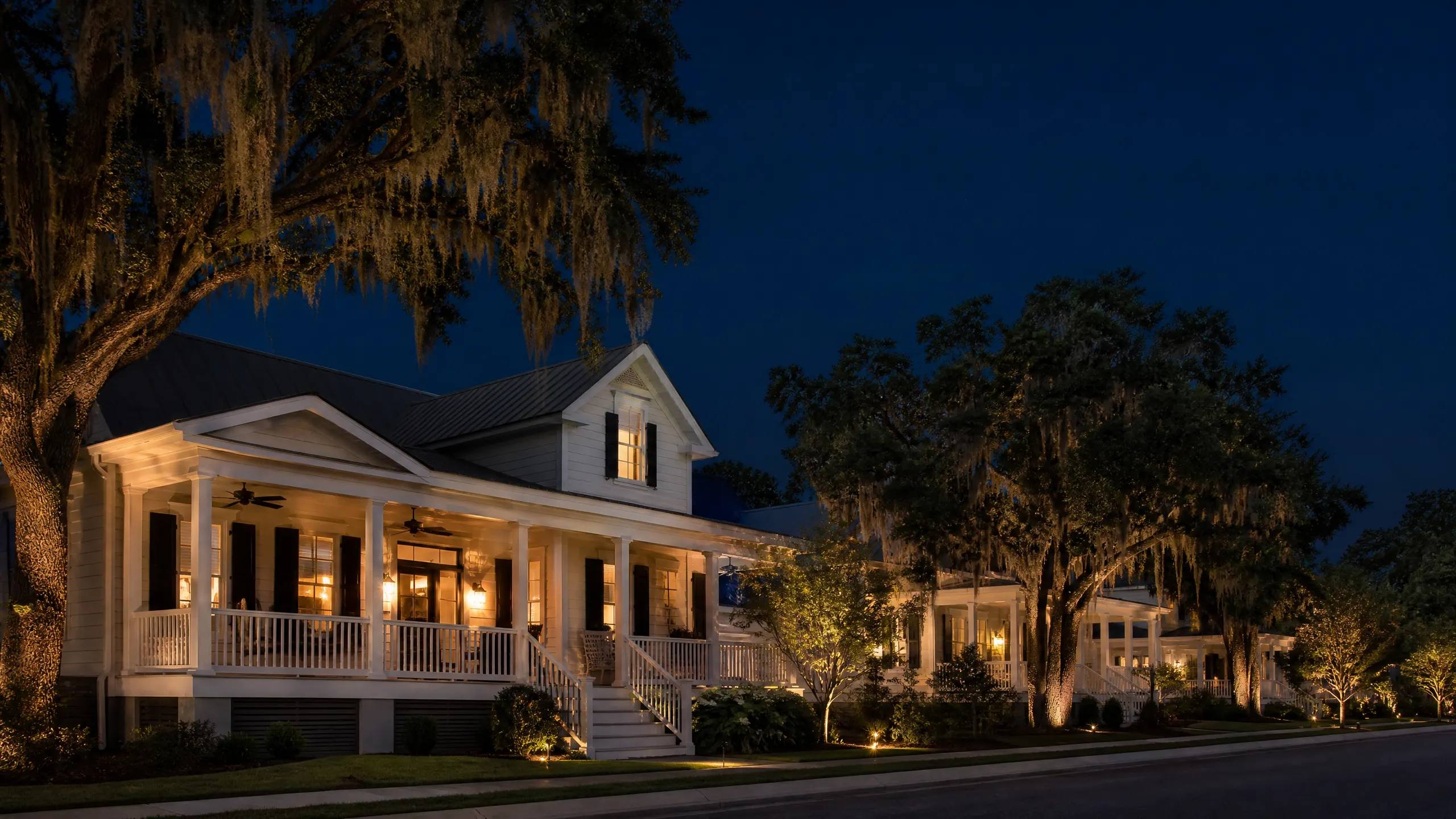 A white Southern-style house with a wraparound porch photographed at night, surrounded by Spanish moss-draped oak trees lit by landscape lights.