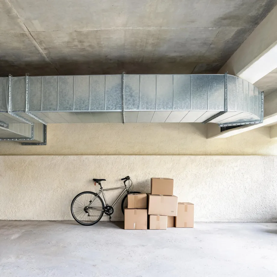 A bicycle leans against a textured beige interior wall next to a stack of five cardboard boxes, beneath a large galvanized metal ventilation duct running along a concrete ceiling.