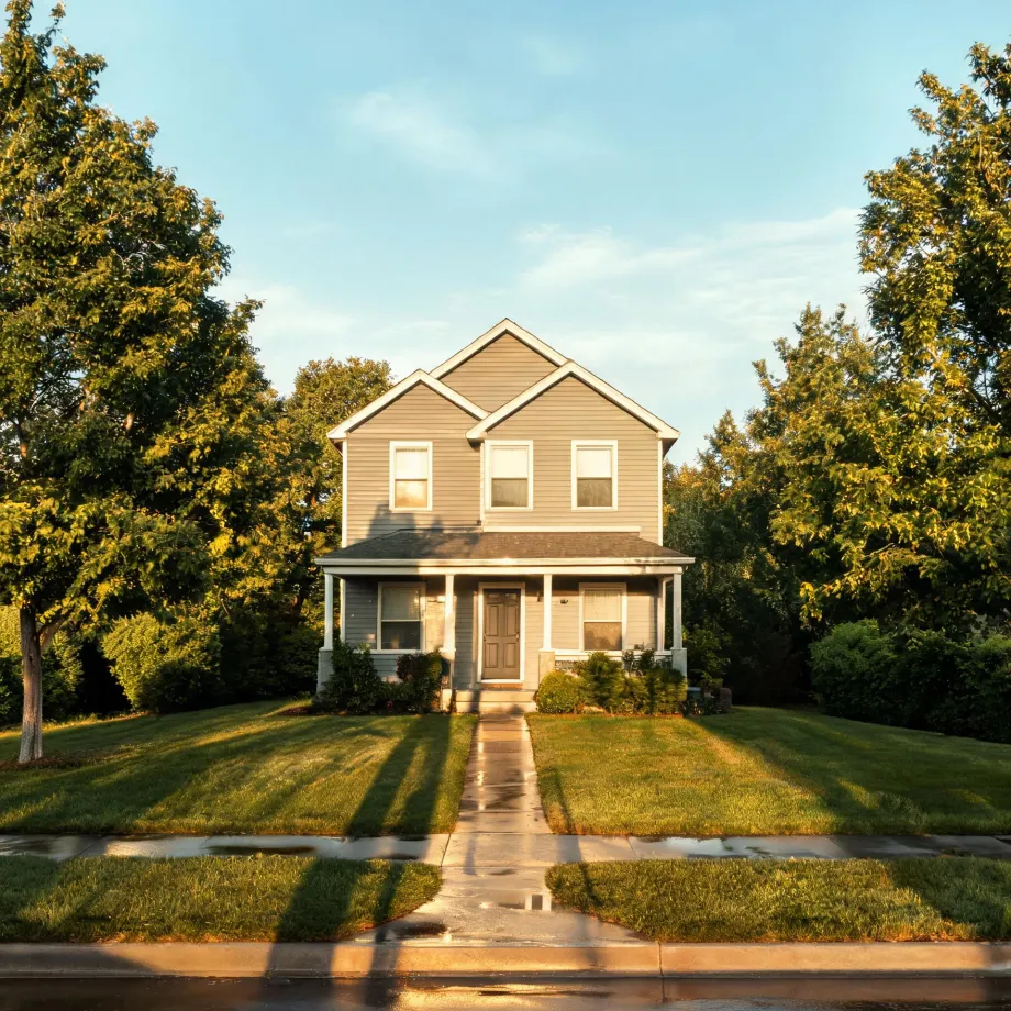 A two-story house with light siding and a covered front porch, fronted by a lawn, a straight concrete walkway to the sidewalk, and large leafy trees on both sides under a clear sky.