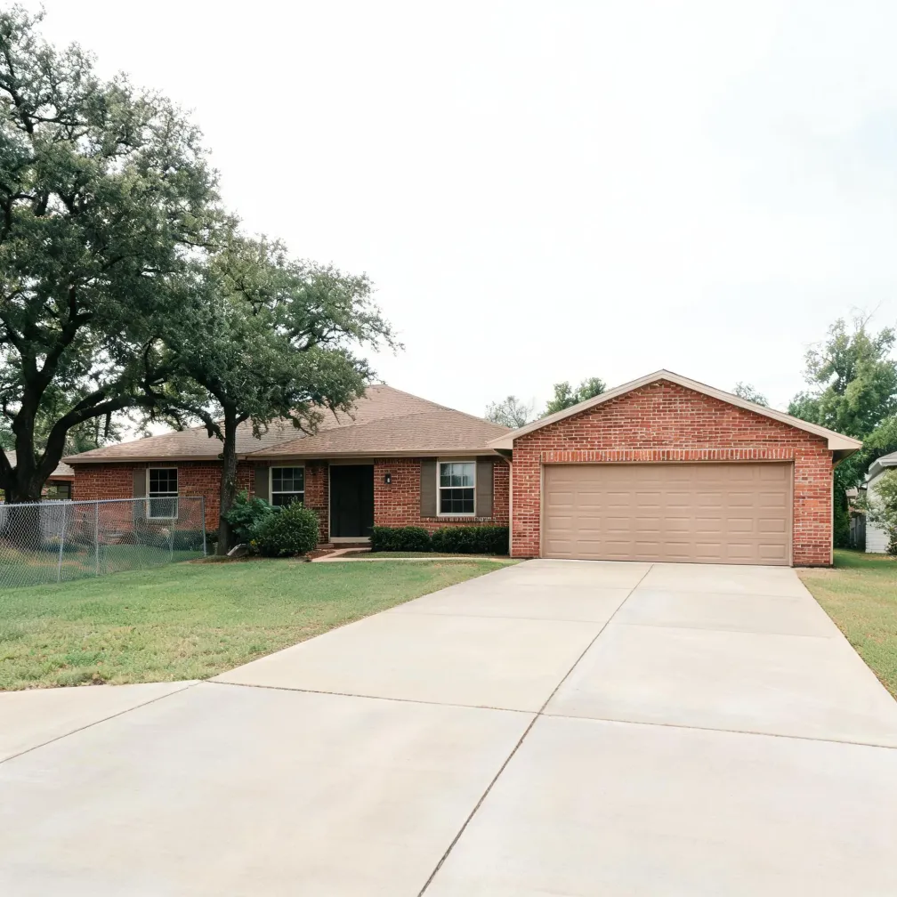 Single-story red brick house with a brown shingled roof, attached two-car garage with a tan door, wide concrete driveway, mowed lawn, chain-link fence at left, and large trees behind the house under an overcast sky.