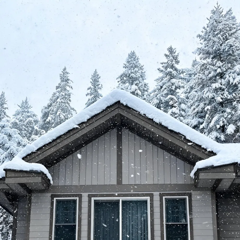 The snow-covered gable roof and gray board-and-batten upper facade of a house with three windows, framed by snow-laden conifers under a pale sky during falling snow.