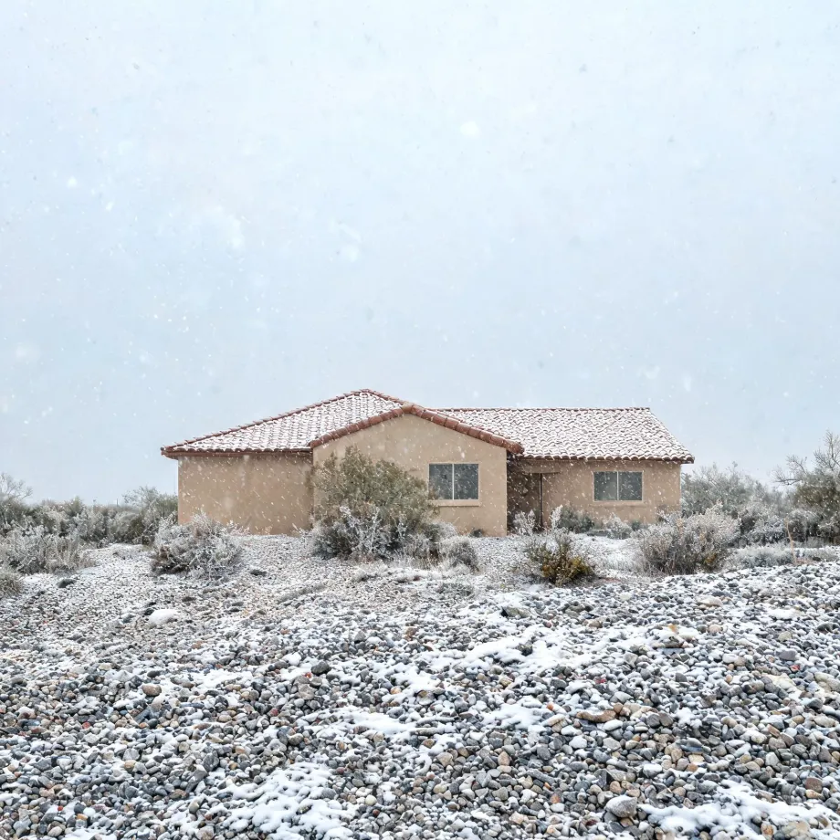 A single-story stucco house with a tile roof stands behind snow-dusted desert scrub and a broad field of gravel during falling snow under a pale sky.