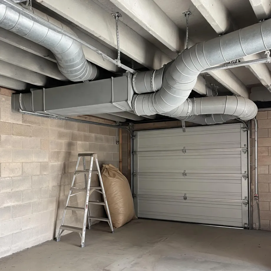 Interior of a garage with concrete block walls, a sectional overhead door, a step ladder and a filled sack, below a ceiling with exposed round and rectangular metal ductwork.