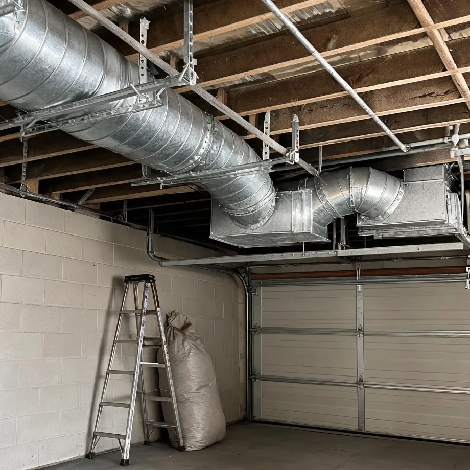 Garage interior with large galvanized round and rectangular ductwork suspended from exposed wood ceiling joists, a concrete block wall, a folding step ladder and a large fabric sack on the floor, and a sectional garage door on the right.