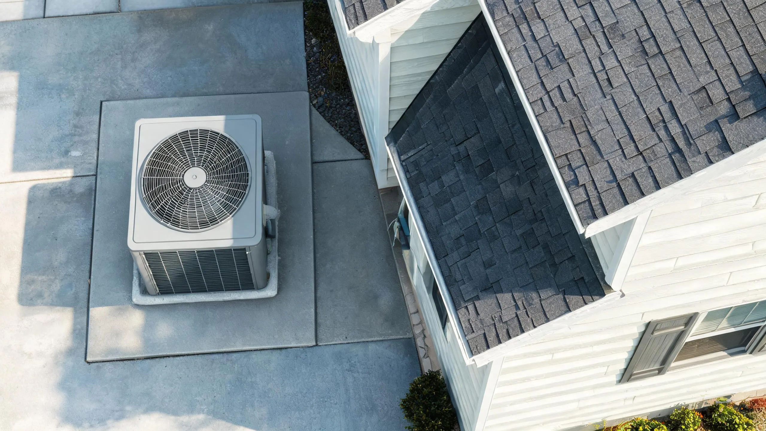 Overhead view of an outdoor AC condenser unit on a concrete pad next to a house with a dark shingle roof.