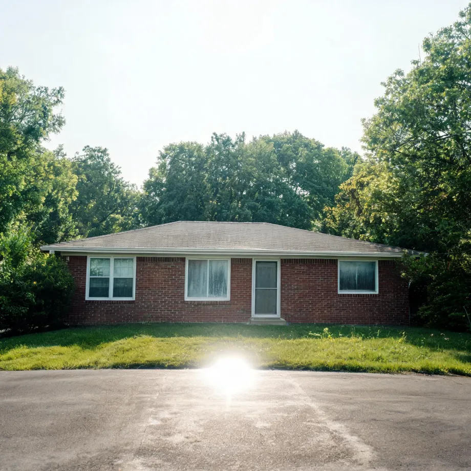 A single-story red brick house with a shingled hip roof, three windows and a central storm door, fronted by a mowed lawn and an asphalt driveway, with dense green trees behind it and sun flare in the foreground.