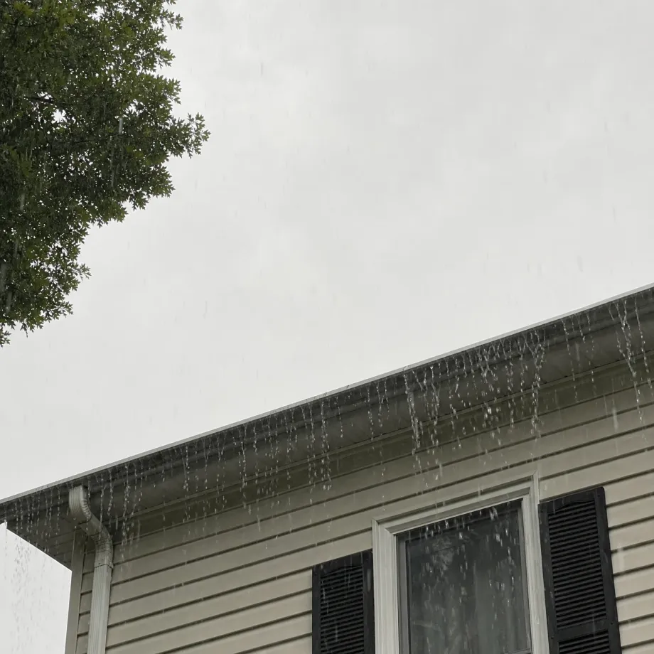 Rain pours off the edge of a gutter along the roofline of a house with beige vinyl siding, a window with dark shutters, and a downspout, with tree branches at the upper left against an overcast sky.
